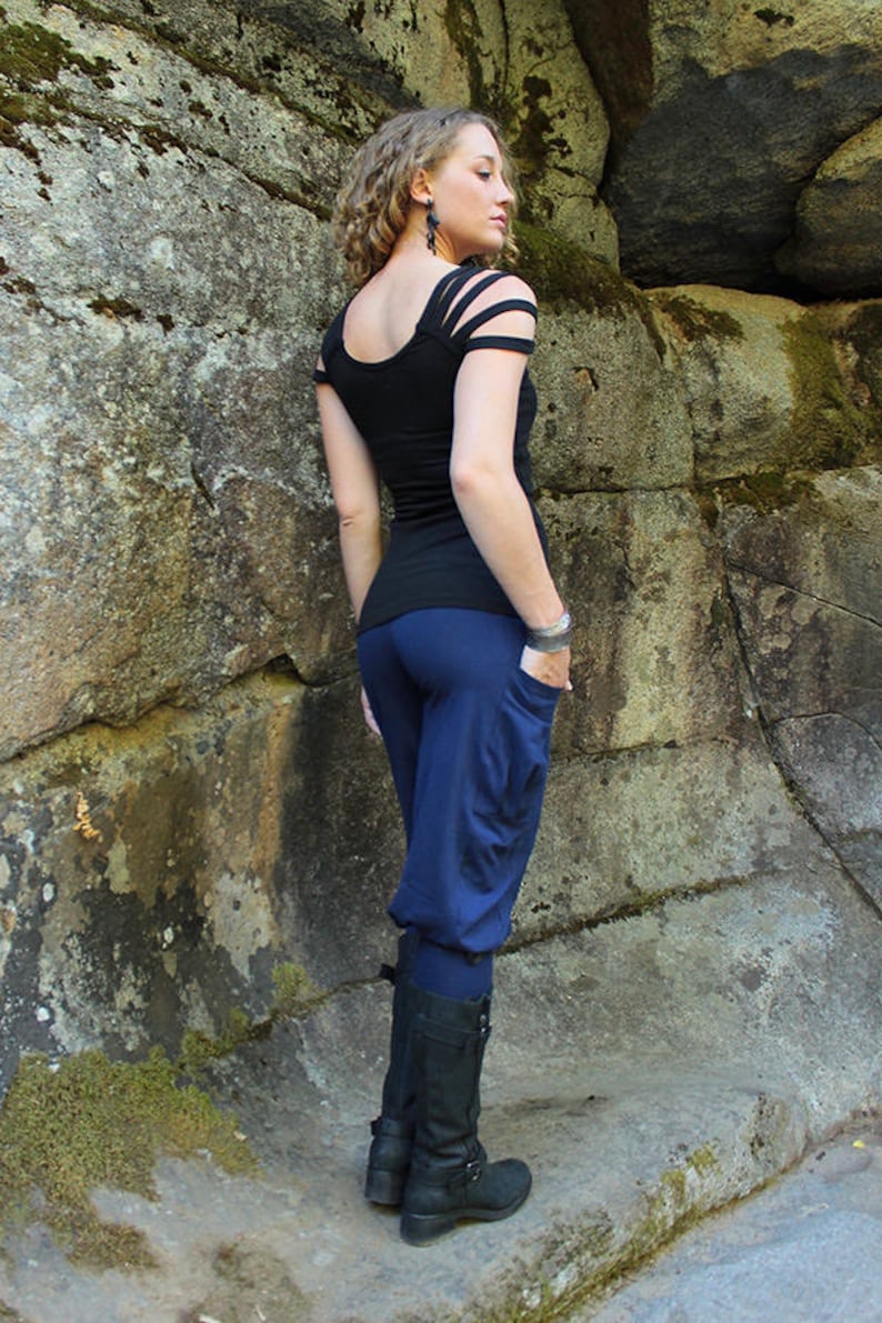 May include: A woman wearing a black top with cut-out shoulders and blue pants. She is standing in front of a rocky wall and wearing black boots.
