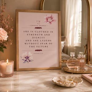 Könnte beinhalten: Ein gerahmter Wandkunstdruck mit hellrosa Hintergrund und dem Text: She is clothed in strength and dignity and she laughs without fear of the future. Proverbs 31:25. Der Druck ist mit rosa und violetten Blumenillustrationen verziert.
