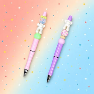 May include: Two ballpoint pens with metallic silver tips and colorful barrels. The left pen has a light pink barrel and a topper featuring a white bear, a green bead, and a translucent bead. The right pen has a purple barrel and a topper with a white bear, a pink leopard print bead, and a light purple bead.