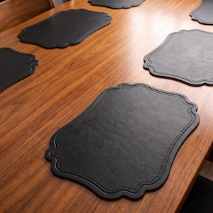 May include: A set of black placemats with a scalloped edge and a decorative stitched border. Each placemat features a smooth, leather-like texture and a distinctively shaped outline, arranged across a wooden surface for dining table settings.