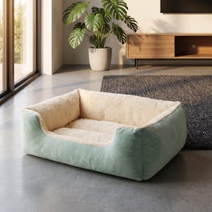 Puede incluir: Una cama para mascotas de forma rectangular con un forro interior de felpa suave en color crema y un tejido exterior liso en verde salvia. La cama cuenta con bordes elevados para mayor soporte y una base acolchada para el descanso.