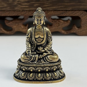 Puede incluir: Una pequeña figura de Buda de metal color dorado en posición de meditación sentada. La figura presenta túnicas detalladas, un ushnisha texturizado y descansa sobre una base de flor de loto estilizada con intrincados pétalos tallados y acabado.