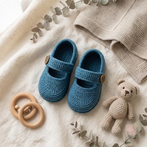 May include: A pair of blue crocheted baby shoes featuring a rounded toe, a single strap across the top secured with a small brown wooden button, and a textured yarn construction. The shoes are accompanied by two smooth wooden rings and a small crocheted bear.