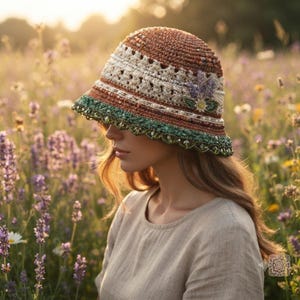 May include: A crochet bucket hat featuring a rust-colored crown, a cream-colored textured band, and a green scalloped brim adorned with beads. The hat is decorated with a small floral appliqué consisting of purple lavender sprigs and a white flower.
