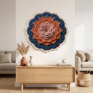 Peut inclure: Une décoration murale circulaire composée de pétales en crochet superposés dans des tons rose poudré, orange brûlé et bleu marine profond. Le motif présente une bordure festonnée crème ornée de petits clous métalliques disposés en rayon.