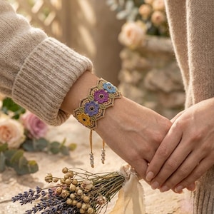May include: A wide, intricate bracelet featuring a series of crocheted floral motifs in yellow, purple, and blue, accented with small beads and green leaf-shaped details. The bracelet has two dangling beaded strands and a scalloped edge design.