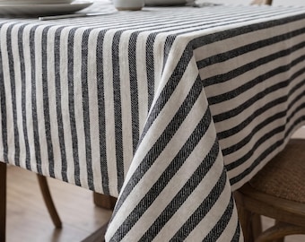 May include: A rectangular tablecloth featuring a repeating pattern of vertical black and off-white stripes. The fabric drapes over the edges of a wooden table, covering the surface completely with its textured, woven appearance.