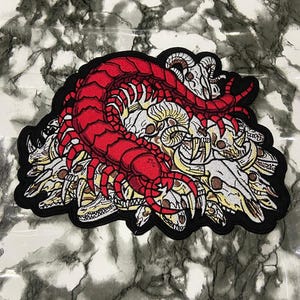 May include: A large embroidered patch featuring a vibrant red centipede coiled atop a cluster of white and beige animal skulls. The design is outlined in a thick black border, showcasing detailed stitching of the insect's segments and the various skull shapes.