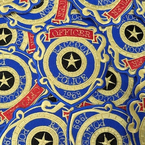 May include: A collection of embroidered patches featuring a blue and gold shield design. Each patch displays a central star, the text RACCOON POLICE, the year 1969, and a red banner with the word OFFICER. The patches are arranged in a layered pattern.