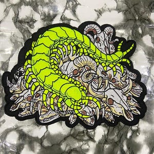 May include: A large embroidered patch featuring a bright neon green centipede coiled over a cluster of white and tan animal skulls. The design is outlined with a thick black border, showcasing detailed stitching of the insect and various skeletal remains.