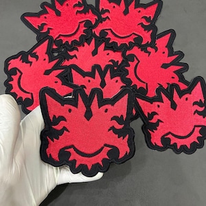 May include: A collection of embroidered patches featuring a stylized red creature design with black outlines. Each patch has a jagged, symmetrical shape with pointed ears and a curved bottom edge, showcasing dense red thread work and black borders.