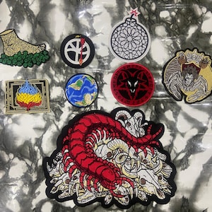 May include: A collection of embroidered patches featuring various designs including a red centipede on a pile of skulls, a goat head, a globe, a bomb, a book with flames, a circular emblem with a red goat, and a foot resting on green leaves.