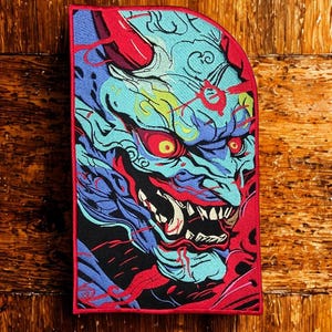 May include: A rectangular embroidered patch featuring a vibrant illustration of a blue-skinned oni mask with sharp white teeth, glowing yellow eyes, and red horns. The design is accented with swirling blue and red patterns on a black background.