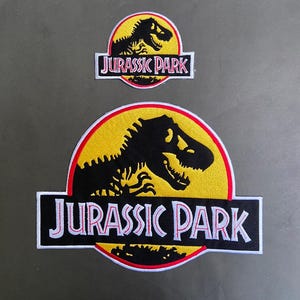 May include: Two embroidered patches featuring the Jurassic Park logo. Each patch displays a black silhouette of a Tyrannosaurus Rex skeleton against a yellow circular background with a red border, and the text JURASSIC PARK in white letters.