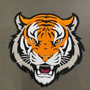 May include: A large embroidered patch featuring the head of a tiger with orange, white, and black fur. The tiger has a fierce expression with an open mouth showing teeth, a dark red tongue, and piercing orange eyes, all outlined in black thread.