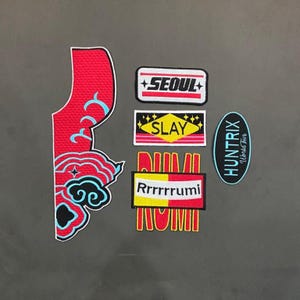 Puede incluir: Una colección de parches bordados que incluye una pieza grande en forma de nube roja y azul, un parche rectangular con la palabra SEOUL, un parche SLAY, un parche Rrrrrrumi RUMI y un parche ovalado con la palabra HUNTRIX World tour.