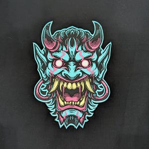 May include: A fabric patch featuring a stylized Hannya mask design in vibrant cyan, pink, and yellow colors. The mask displays sharp horns, glowing eyes, and prominent fangs, with a black background and detailed linework throughout the graphic.
