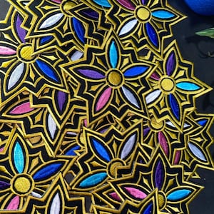 May include: A collection of star-shaped embroidered patches featuring a central gold circle surrounded by five petals in alternating colors of blue, purple, pink, and white, all outlined with thick black and gold thread borders.