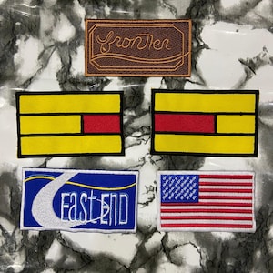 May include: Five rectangular embroidered patches. The top patch is brown with the word frontier. Two middle patches feature yellow and red horizontal stripes. The bottom left patch is blue with a white crescent and the text EAST END. The bottom right is a US flag.