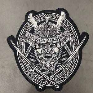 Puede incluir: Un parche bordado en blanco y negro que presenta una máscara de samurái detallada con un casco tradicional. El diseño está encerrado en un borde circular con un patrón trenzado, y dos espadas cruzadas se sitúan frente a la máscara.