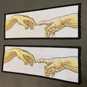 May include: Two rectangular embroidered patches featuring a reproduction of the Creation of Adam hands. Each patch has a white background, tan skin-toned hands reaching toward each other, and a thick black stitched border framing the design.
