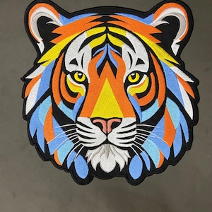 May include: An embroidered patch featuring a stylized tiger head with vibrant colors including orange, yellow, light blue, and white. The design is outlined in black thread, showcasing detailed stitching of the tiger's facial features and fur.