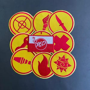 May include: A collection of circular embroidered patches in yellow with red borders and symbols, including a knife, flame, wrench, and mace. A rectangular patch featuring a red and white flag with a bomb icon and the text RED is centered.