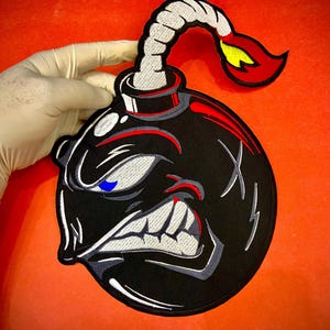 May include: An embroidered patch featuring a black cartoon bomb with an angry facial expression, white teeth, and a single blue eye. The bomb has a lit fuse with a red and yellow flame at the top, all outlined in black thread on a black fabric base.