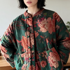 May include: A quilted jacket featuring a dark green base with a large-scale floral pattern in shades of peach and pink. The garment has a mandarin collar, long sleeves, two front patch pockets, and a front closure with small green fabric buttons.