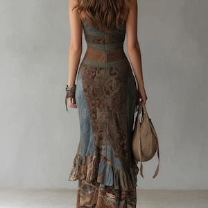 May include: A floor-length sleeveless dress featuring a mix of blue and brown paisley and floral patterns. The garment has a fitted bodice, a defined waistline, and a tiered skirt with a ruffled hem. A woven tan handbag is held at the side.