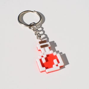 May include: A pixel art style keyring featuring a small bottle shape with a white outline, a bright red liquid centre, and a brown stopper. The charm is attached to a silver metal split ring by a short silver chain, creating a retro gaming aesthetic.