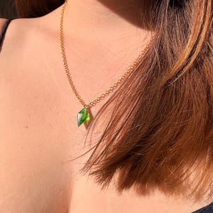 Puede incluir: Un collar de cadena de metal dorado con un pequeño colgante de cristal verde translúcido y facetado en forma de diamante. El colgante cuelga de una cadena de eslabones sencilla, captando la luz para mostrar su vibrante tono verde esmeralda.