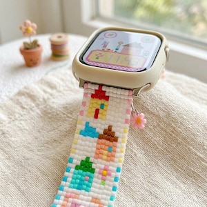 May include: A smartwatch with a cream-colored protective case and a white beaded watch band featuring a repeating pattern of small colorful houses in yellow, orange, blue, and pink. A small pink flower charm hangs from the side of the watch band.
