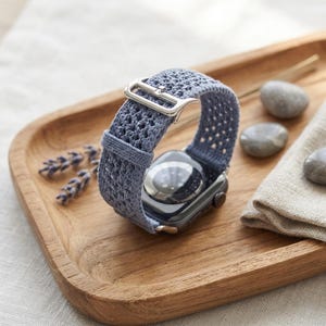 Peut inclure: Une montre connectée dotée d'un bracelet en crochet bleu pervenche texturé avec une boucle en métal argenté. Le boîtier de la montre est gris foncé avec un capteur circulaire visible sur le dessous, présentant un motif tissé détaillé.