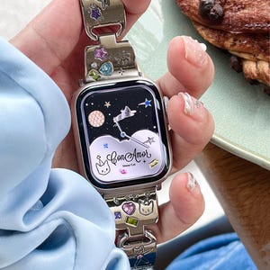 May include: A silver-toned smartwatch strap featuring embedded colourful crystals, star-shaped accents, and engraved cat motifs. The watch face displays a night sky graphic with a moon, stars, clouds, and the text Con Amor Moon Cat in script.