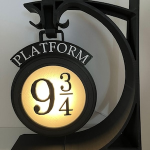 May include: A black wall-mounted lamp featuring a circular sign that reads PLATFORM 9 3/4. The sign is illuminated from within, casting a warm glow through the center. The structure is supported by a curved black arm and a flat base.