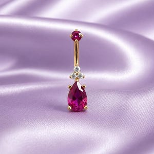 May include: A gold navel piercing jewelry piece featuring a round, deep pink gemstone at the top, a cluster of three small clear stones in the center, and a large, pear-shaped, deep pink gemstone drop at the bottom, all set in a polished gold metal.