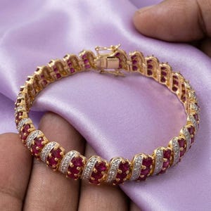 May include: A gold-toned bracelet featuring a repeating pattern of oval-shaped red gemstones alternating with small, clear, sparkling stones. The piece includes a secure box clasp mechanism and is constructed with a series of linked segments.