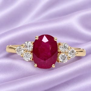 May include: A gold ring featuring a large, oval-cut, deep red ruby at the center. The ruby is flanked on both sides by a cluster of three small, round, brilliant-cut white diamonds set in gold prongs, creating a symmetrical and elegant design.