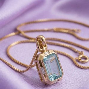 Puede incluir: Un collar de oro con un colgante rectangular que presenta una gran piedra preciosa de color azul claro en talla esmeralda en el centro. El colgante está enmarcado por pequeñas piedras transparentes en talla baguette y cuelga de una cadena.