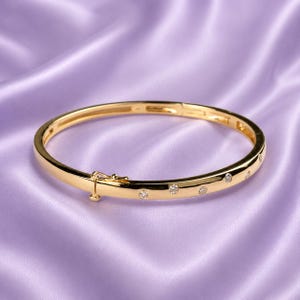 Puede incluir: Una pulsera rígida articulada en tono dorado pulido que presenta una serie de pequeñas piedras redondas y transparentes incrustadas en la banda metálica. La pulsera incluye un cierre integrado seguro en un lado, creando una forma circular.