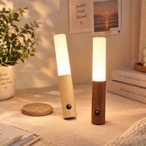 May include: Two cylindrical portable lamps with wooden bases and frosted white light diffusers. Each lamp features a small black circular motion sensor button on the front. One lamp has a light oak finish, while the other has a dark walnut finish.