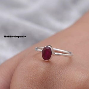 Könnte beinhalten: Ein Silberring mit einem einzelnen, oval geschliffenen, tiefroten Rubin, der in eine schlichte Metallschiene gefasst ist. Der Text TheSilverEmpireCo ist oben links im Bild zu sehen. Der Ring wird an einem Finger vor grauem Hintergrund gezeigt.
