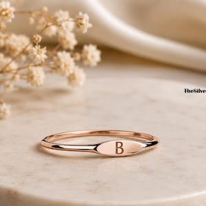 Puede incluir: Un anillo de tono oro rosa con una banda fina y pulida y una pequeña placa ovalada horizontal grabada con la letra B. El texto TheSilverEmpireCo es visible en la esquina superior derecha de la imagen.