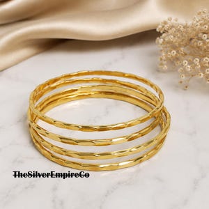 Puede incluir: Un conjunto de cuatro brazaletes dorados con una superficie martillada y texturizada. Las pulseras son circulares y tienen un acabado metálico reflectante. El texto TheSilverEmpireCo está impreso en negro en la esquina inferior izquierda.