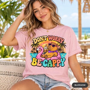 Woman in sunglasses at a beach bar wearing the "Don't Worry Be Cappy" capybara beach T-shirt by SarcasmArt in Blossom (soft pink).
