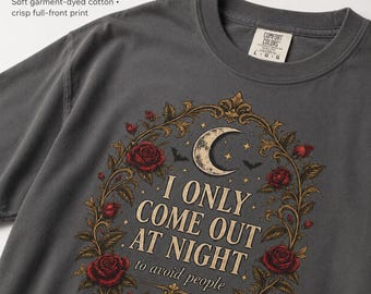 Op de afbeelding: Een antracietgrijs katoenen t-shirt met een grafische print van een maansikkel, vleermuizen en rode rozen in een sierlijk gouden kader. De tekst luidt I ONLY COME OUT AT NIGHT to avoid people. Het shirt heeft een ronde hals en maatlabel.