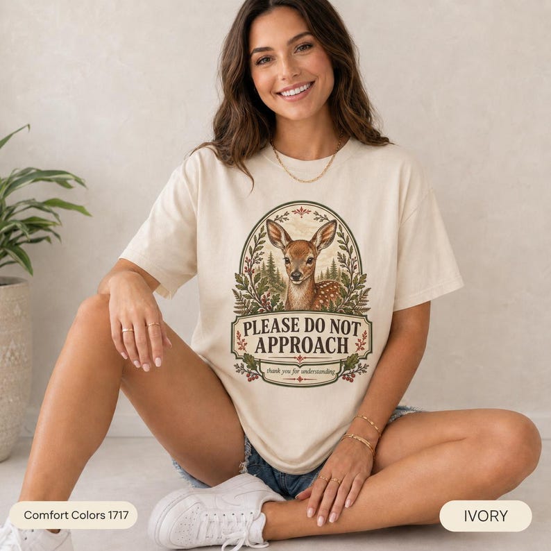 Smiling woman sitting on the floor wearing the "Please Do Not Approach" vintage fawn introvert T-shirt by SarcasmArt in Ivory – a relaxed unisex forestcore tee with a vintage woodland deer fawn in an oval frame of ferns and wildflowers above the words PLEASE DO NOT APPROACH.