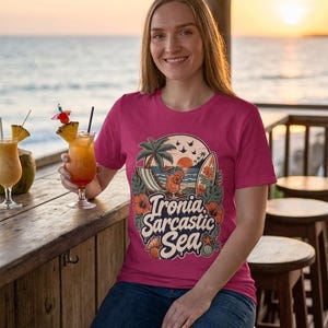 Smiling woman at a beach bar at sunset wearing the "Ironia, Sarcastic Sea" retro beach T-shirt by SarcasmArt in raspberry pink – an anti-social vacation tee with a vintage tropical beach scene with palm tree, deck chair, cocktail, surfboard, sunset, hibiscus flowers and shells around the script Ironia, Sarcastic Sea.