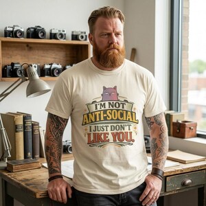 Bearded man in a cozy study wearing the "I'm Not Anti-Social, I Just Don't Like You" sarcastic introvert T-shirt by SarcasmArt in Ivory.